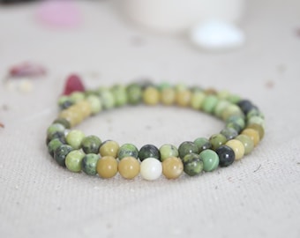 May include: A beaded bracelet composed of small, round, polished stones in various shades of olive green, lime green, and dark forest green, featuring a single smooth, opaque white bead positioned near the front of the circular arrangement.