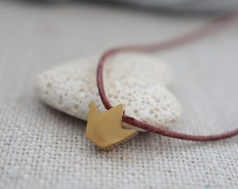 May include: A gold-colored pendant in the shape of a crown is strung on a thin, reddish-brown cord. The necklace rests on a textured, off-white stone against a neutral background. The crown pendant is a simple, geometric design.