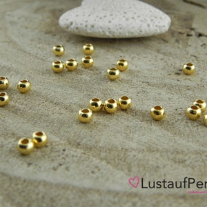 May include: A close-up of a group of 20 gold-colored round beads scattered on a light brown wooden surface. The beads are smooth and shiny.