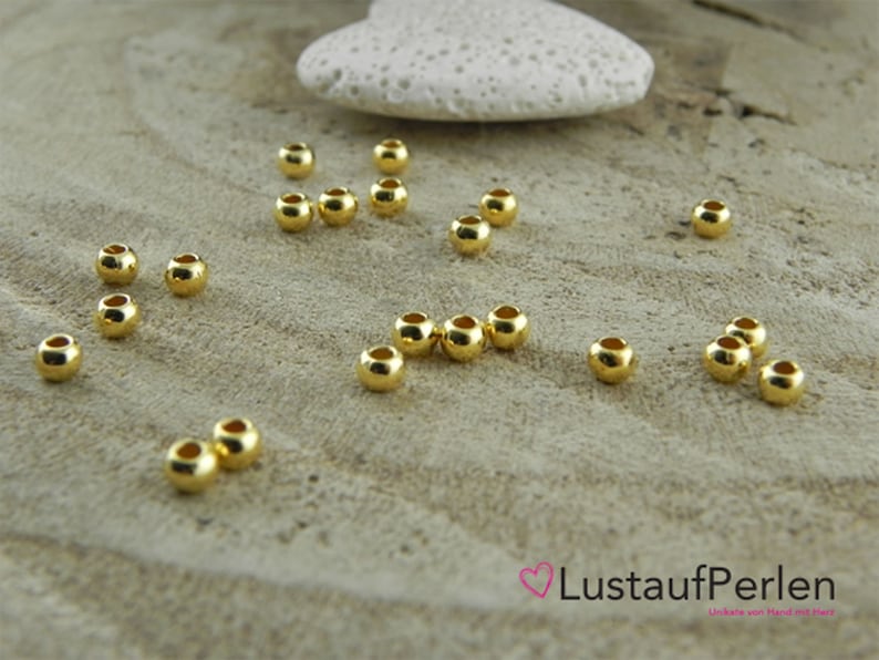May include: A close-up of a group of 20 gold-colored round beads scattered on a light brown wooden surface. The beads are smooth and shiny.