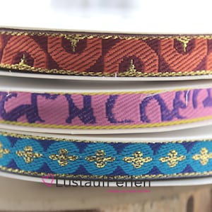 Könnte beinhalten: Drei Rollen farbiges, gemustertes Band mit goldenen Akzenten. Das obere Band ist braun mit einem goldenen Muster, das mittlere Band ist rosa mit einem violetten Muster und das untere Band ist blau mit einem goldenen Muster.