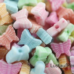 Könnte beinhalten: Nahaufnahme von bunten Schmetterlingsperlen in Pastellfarben. Die Perlen sind in Rosa, Blau, Grün und Orange gehalten. Jede Perle hat ein Loch zum Auffädeln. Der Text "Lust auf Perlen" ist sichtbar.