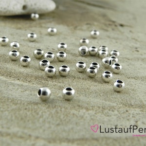 May include: A close-up of a group of silver round beads scattered on a light brown surface. The beads are smooth and shiny.