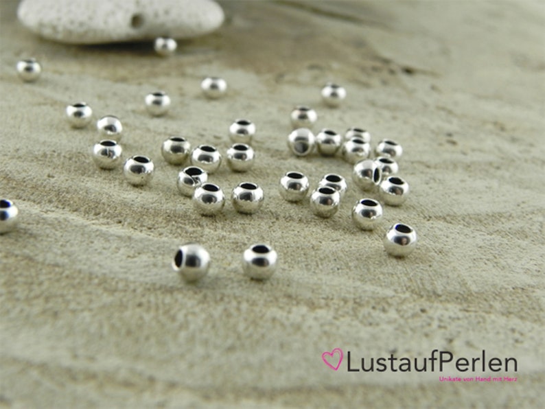 May include: A close-up of a group of silver round beads scattered on a light brown surface. The beads are smooth and shiny.