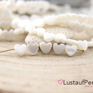 Könnte beinhalten: Nahaufnahme von weißen herzförmigen Perlen, die auf einer goldfarbenen Nadel aufgereiht sind. Im Hintergrund befinden sich mehrere Stränge ähnlicher Perlen sowie ein Logo. Die Perlen haben einen perlmuttartigen Glanz und sind auf einer hellen Holzoberfläche angeordnet.