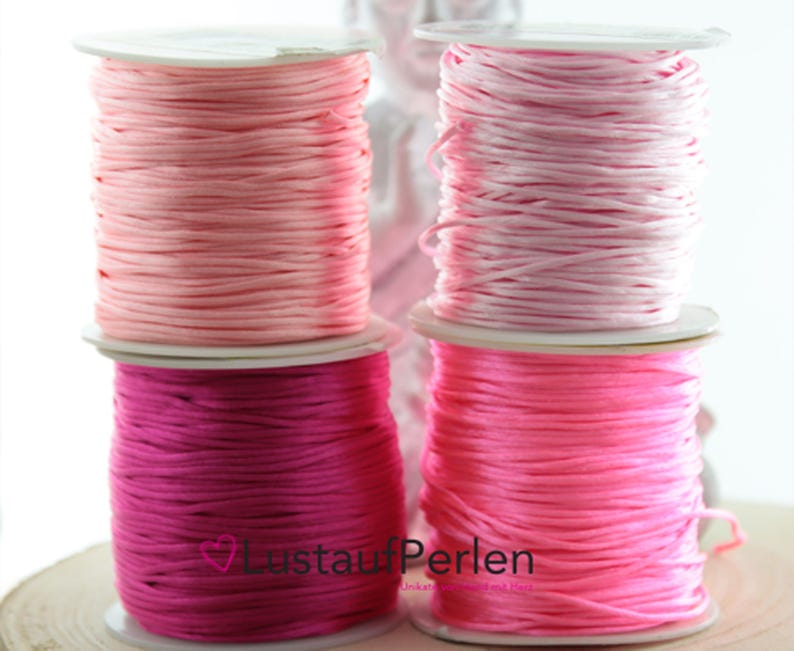 May include: Four spools of waxed cord in pink, light pink, hot pink, and a darker shade of pink. The spools are labeled "LustaufPerlen".