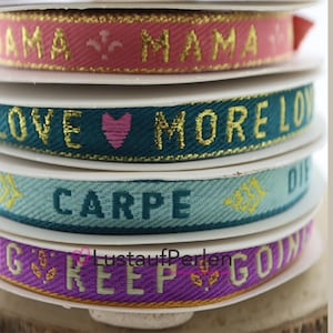 Könnte beinhalten: Vier Spulen mit Band mit goldener Einfassung und verschiedenen Farben und Sprüchen. Die oberste Spule ist rosa mit goldenen Buchstaben, die "Mama Mama" sagen. Die zweite Spule ist türkis mit goldenen Buchstaben, die "I Love More Love" sagen. Die dritte Spule ist hellblau mit goldenen Buchstaben, die "Carpe Diem" sagen. Die unterste Spule ist lila mit goldenen Buchstaben, die "Keep Going" sagen.