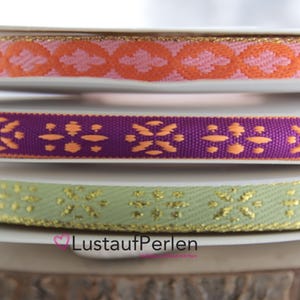 Könnte beinhalten: Drei Rollen gemustertes Band mit goldenen Akzenten. Das obere Band ist rosa mit orangefarbenen Akzenten, das mittlere Band ist lila mit orangefarbenen Akzenten und das untere Band ist grün mit goldenen Akzenten.