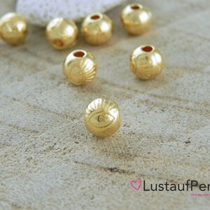 May include: Gold-colored round beads with an eye design. The beads have a hole in the center for stringing.