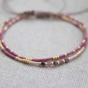 Könnte beinhalten: Ein zartes, zweireihiges Armband mit kleinen malvenfarbenen und hellrosa Saatperlen, die mit winzigen metallisch goldenen Zwischenperlen durchsetzt sind. Die untere Reihe enthält eine Serie kleiner, facettierter Edelsteinperlen.