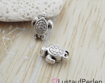 May include: Two small, silver-colored turtle charms. The charms have detailed shells and flippers. The image is on a light-colored wooden surface. The charms are likely for jewelry making or crafting.