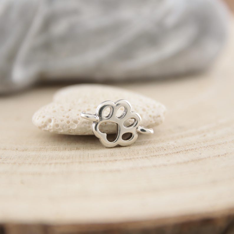 May include: Silver charm in the shape of a paw print. The charm has a small loop at the top for attaching to a bracelet or necklace.