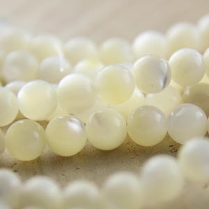 Könnte beinhalten: Nahaufnahme von cremeweißen, runden Perlen. Die Perlen haben einen perlmuttartigen Glanz und sind aufgefädelt, was darauf hindeutet, dass sie für die Schmuckherstellung bestimmt sind. Die Perlen sind glatt und gleichmäßig groß, mit einer leichten Farbvariation.