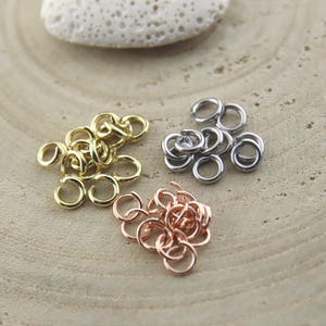 Könnte beinhalten: Eine Ansammlung kleiner Metall-Biegeringe, die in drei getrennten Gruppen angeordnet sind. Die Sets bestehen aus goldfarbenen, silberfarbenen und roségoldfarbenen kreisförmigen Ringen, die jeweils eine kleine Öffnung zum Verbinden von Schmuckteilen aufweisen.