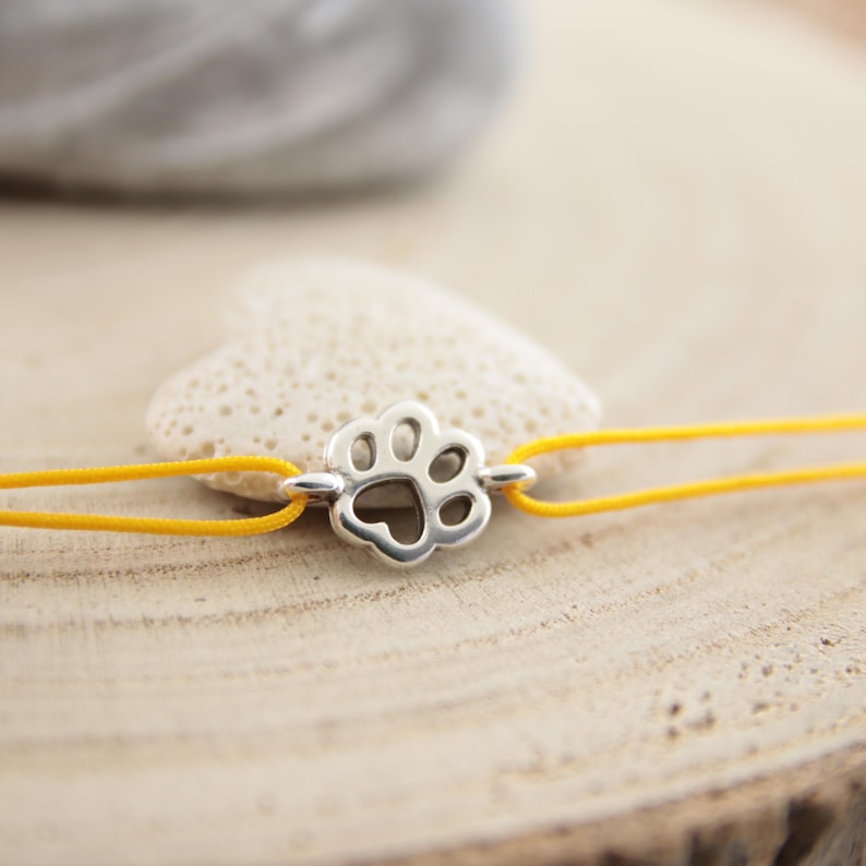 May include: A yellow string bracelet with a silver paw print charm. The charm has a heart-shaped cutout in the centre.