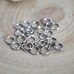 Könnte beinhalten: Eine Ansammlung kleiner, kreisförmiger Biegeringe aus silberfarbenem Metall. Jeder einzelne Ring weist einen kleinen Schlitz auf, wodurch sie sich öffnen und schließen lassen, um Schmuckkomponenten oder andere Bastelartikel zu verbinden.