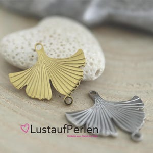 Könnte beinhalten: Zwei Ginkgo-Blatt-Anhänger, einer in Goldoptik und einer in Silberoptik. Jeder Metallanhänger hat ein fächerförmiges Design mit feinen linearen Details und zwei kleinen kreisförmigen Ösen zur Befestigung, die oben und unten angebracht sind.