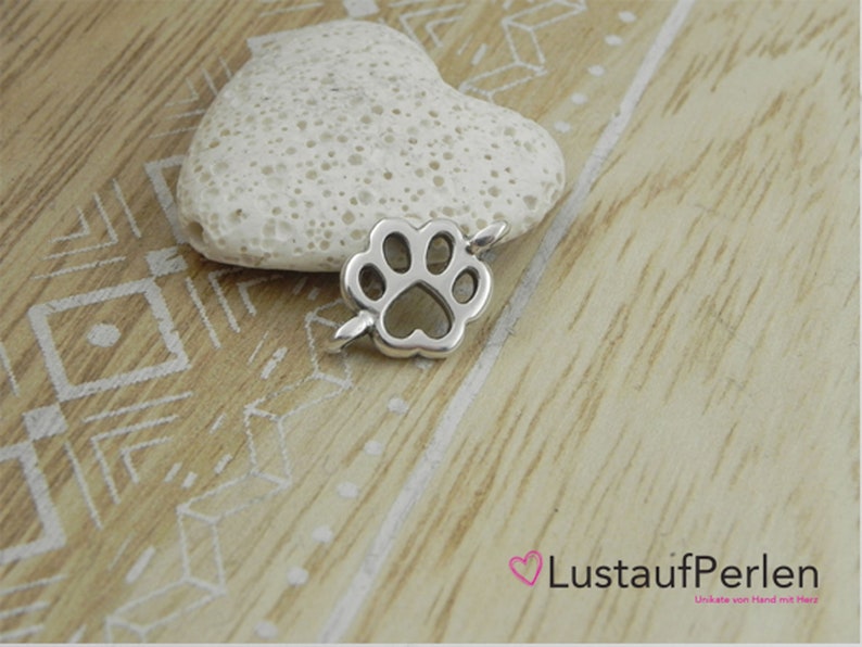 May include: Silver charm in the shape of a paw print. The charm is on a white heart-shaped rock.