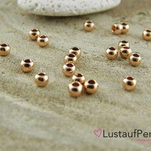 May include: A close-up of a group of 20 gold-colored round beads. The beads are scattered on a light brown surface.