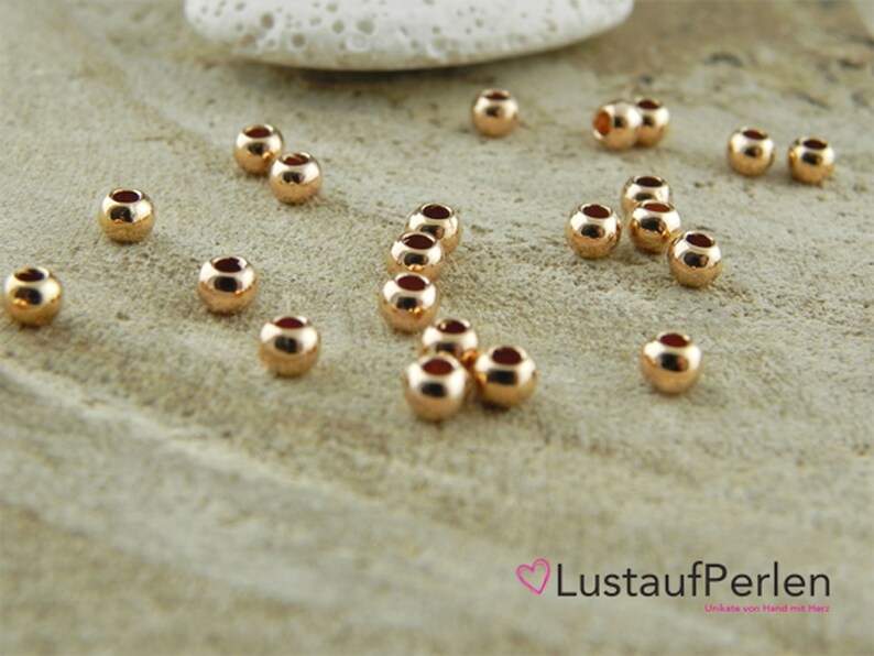 May include: A close-up of a group of 20 gold-colored round beads. The beads are scattered on a light brown surface.