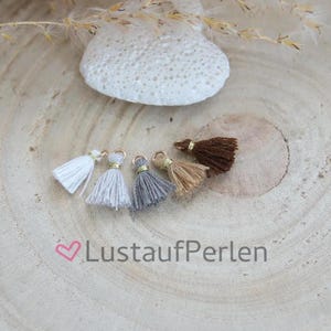 以下が含まれることがあります： 白、ライトグレー、ダークグレー、ベージュ、ダークブラウンの5つの小さな糸のタッセル。それぞれの上部にはゴールドトーンの金属製丸カンが付いています。画像の下部には、ピンクのハートアイコンが付いた「LustaufPerlen」というテキストが印刷されています。