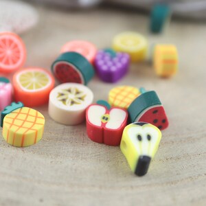 Könnte beinhalten: Eine Sammlung farbenfroher, fruchtförmiger Perlen, darunter Orangenscheiben, Wassermelonen, Trauben und Ananas. Die Perlen bestehen aus einem glatten Material und sind hell gefärbt, mit detaillierten Designs. Ideal für Schmuckherstellung und Basteln.