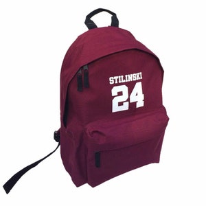 Op de afbeelding: Bordeauxrode rugzak met zwarte banden en een witte rits. De rugzak heeft een witte patch met de tekst "STILINSKI 24" op de voorkant.