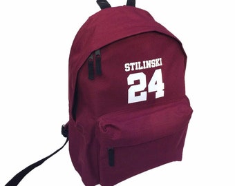 May include: Burgundy red backpack with black straps and a white zipper. The backpack has a white patch with the text "STILINSKI 24" on the front.