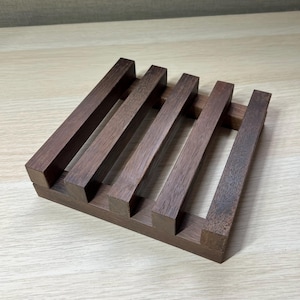 Könnte beinhalten: Ein quadratischer Untersetzer aus dunkelbraunem Holz mit fünf erhöhten Leisten. Der Untersetzer ist aus Naturholz gefertigt und hat einen rustikalen Look.
