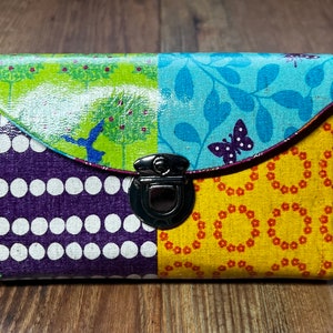 Könnte beinhalten: Eine bunte Patchwork-Geldbörse mit einem silbernen Verschluss. Die Geldbörse hat einen grünen, blauen, violetten und gelben Stoff mit verschiedenen Mustern.