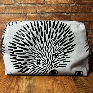 Könnte beinhalten: Eine rechteckige, hellgraue Kosmetiktasche mit einem großen, schwarz-weißen Igel-Design. Der Igel hat ein detailliertes Gesicht mit runden Augen und einer schwarzen Nase. Die Tasche ist aus Stoff und steht auf einer Holzoberfläche.