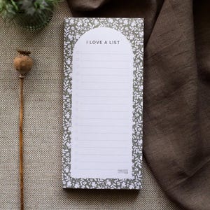 May include: A grey notepad with a white floral border and the text "I love a list" on the front. The notepad has a white lined interior and is perfect for jotting down notes, to-do lists, or ideas.