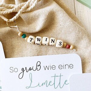 May include: A white card with the text "So gro&szlig; wie eine Limette" in black and green script. A wooden bead necklace with the word "TWINS" spelled out in black letters on white beads.