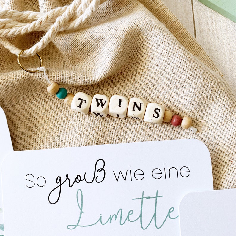 May include: A white card with the text "So gro&szlig; wie eine Limette" in black and green script. A wooden bead necklace with the word "TWINS" spelled out in black letters on white beads.