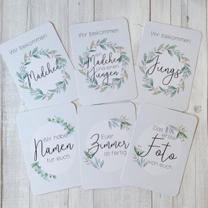 May include: Six white cards with green and pink floral wreaths and black text. The text on the cards reads: "Wir bekommen ein Madchen und einen Jungen", "Wir bekommen Jungs", "Wir bekommen Madchen", "Wir haben Namen fur euch", "Euer Zimmer ist fertig", "Das erste Foto von euch".