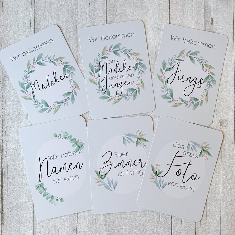 May include: Six white cards with green and pink floral wreaths and black text. The text on the cards reads: "Wir bekommen ein Madchen und einen Jungen", "Wir bekommen Jungs", "Wir bekommen Madchen", "Wir haben Namen fur euch", "Euer Zimmer ist fertig", "Das erste Foto von euch".