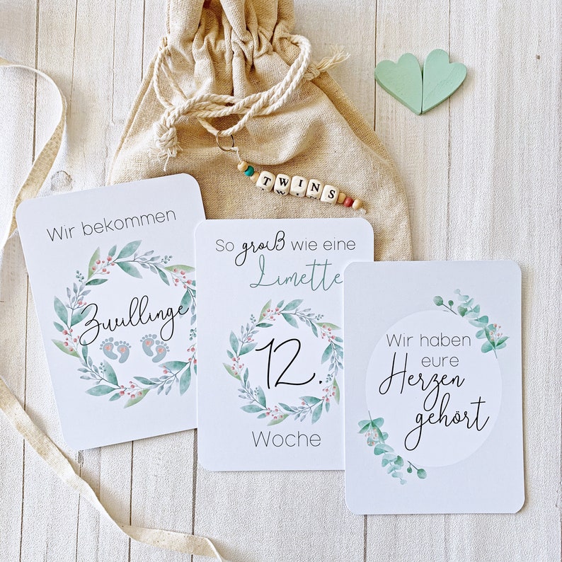 May include: Three white cards with green floral wreaths and text. The first card says "Wir bekommen Zwillinge". The second card says "So gro&szlig; wie eine Limette 12. Woche". The third card says "Wir haben eure Herzen geh&ouml;rt".