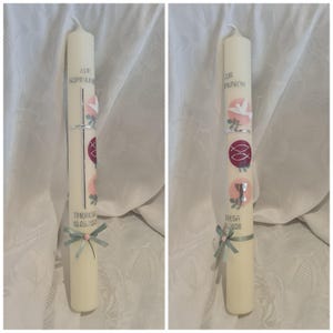 Puede incluir: Una vela crema con símbolos religiosos y diseños florales. La vela tiene texto que dice "ZUR KOMMUNION" y "Theresa 10.05.2020". Una cinta azul claro con una flor rosa adorna la base.