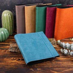Può includere: Una collezione di sei quaderni rettangolari con copertine morbide effetto camoscio in blu, arancione, bordeaux, verde, beige e marrone. Ogni quaderno presenta una chiusura con elastico in tinta e una fodera interna nera, disposti in verticale.
