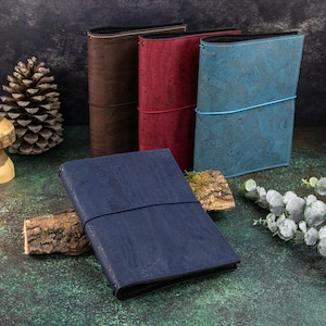 May include: Four rectangular journals with textured cork covers in dark brown, deep red, bright blue, and navy blue. Each journal is secured with a thin, color-coordinated elastic band wrapped horizontally around the center of the cover.