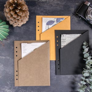 May include: Three rectangular paper pocket folders with six-hole punched edges for binders. One folder is light brown, one is mustard yellow, and one is black. Each folder features a diagonal opening containing printed paper inserts with text.
