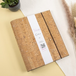 May include: A cork notebook with a white label that says "Valhalla's Order A5" and has a QR code. The notebook has a brown elastic closure.