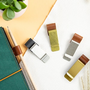 Puede incluir: Cinco clips para cuaderno de cuero y metal en diferentes colores. Los clips están sobre una superficie blanca con un cuaderno verde y un lápiz en el fondo.