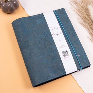 Puede incluir: Un archivador de anillas A5 de color verde azulado con una superficie texturizada, una banda elástica verde azulado y una etiqueta blanca con texto y un código QR. El archivador está sobre una superficie naranja claro.