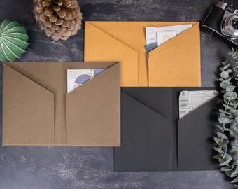 Könnte beinhalten: Drei Papiermappen in Beige, Braun und Schwarz, jeweils mit zwei Innentaschen ausgestattet. Die Mappen enthalten verschiedene Papiere mit gedrucktem Text und Grafiken. Sie sind auf einer dunklen Oberfläche neben einem Tannenzapfen angeordnet.