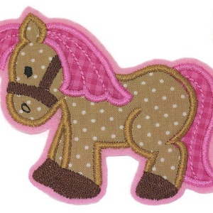 Puede incluir: Una aplicación de caballo marrón con lunares blancos y crin y cola rosas. El caballo tiene una brida marrón y está de pie sobre sus cuatro patas.