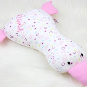 Può includere: Giocattolo di peluche rosa e bianco a forma di anatra con un becco rosa e occhi neri. Il giocattolo ha uno sfondo bianco con pois rosa, viola e verdi e la parola "baby" ricamata sul lato.