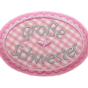 Könnte beinhalten: Rosa Stoffoval mit einem weißen und rosa karierten Muster und dem Text "große Schwester" in silberner Stickerei.
