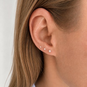 Könnte beinhalten: Drei kleine, runde, weiße Perlenohrstecker, die in einer Reihe entlang des Ohrläppchens angeordnet sind. Jeder Ohrstecker besteht aus einer einzelnen, glatten, kugelförmigen Perle auf einem Metallstift, was ein einheitliches Erscheinungsbild ergibt.