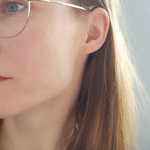 Pu&ograve; includere: Primo piano di un orecchio con un piccolo orecchino a cerchio d'oro. La persona indossa degli occhiali e ha i capelli lunghi e castani.